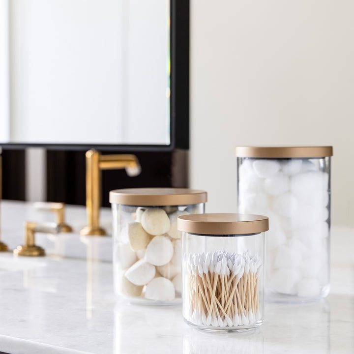clear canisters with gold lids organizing bathroom items like cotton balls, q-tips and face sponges 